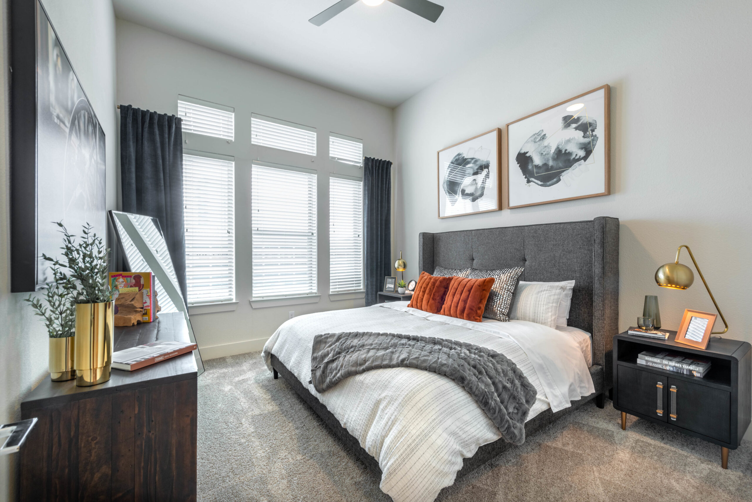 Model bedroom at our Fort Worth apartments, featuring grey carpets, abstract wall art, and three large windows.